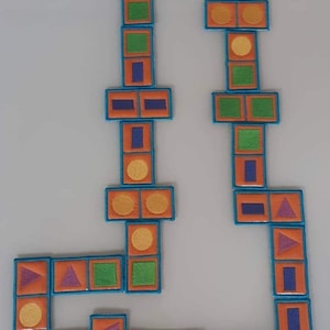 Puede incluir: Un juego de dominó de colores con varias formas y colores, incluyendo naranja, verde, azul, amarillo y morado. Los dominós están dispuestos en un patrón sobre una superficie gris claro.