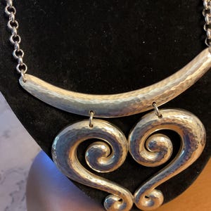 Puede incluir: Collar plateado con acabado martillado. El collar presenta una barra curva en la parte superior y dos colgantes grandes en forma de coraz&oacute;n. La cadena es de estilo eslab&oacute;n simple.