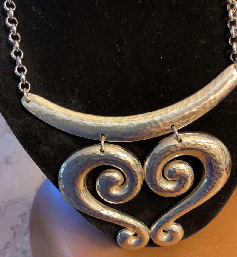 Puede incluir: Collar plateado con acabado martillado. El collar presenta una barra curva en la parte superior y dos colgantes grandes en forma de coraz&oacute;n. La cadena es de estilo eslab&oacute;n simple.