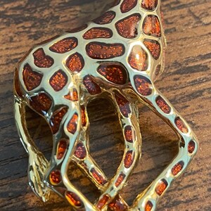 May include: A gold-toned giraffe brooch with a detailed design. The giraffe has a brown and orange spotted pattern on its body and a small green gem for an eye. The brooch is set against a wooden surface.