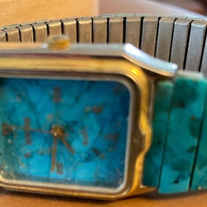 May include: A rectangular wristwatch with a gold-colored case and a turquoise face. The watch has a silver-colored, expandable band and turquoise-colored links. The watch hands are gold-colored.