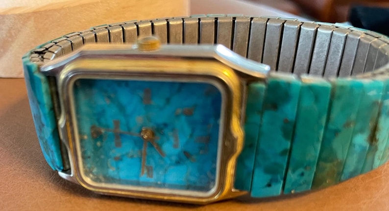 May include: A rectangular wristwatch with a gold-colored case and a turquoise face. The watch has a silver-colored, expandable band and turquoise-colored links. The watch hands are gold-colored.