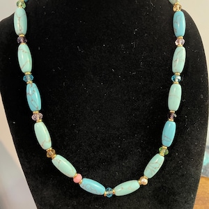 May include: A turquoise beaded necklace with oval-shaped beads. The necklace features gold-colored spacers and accents of pink, purple, and blue beads. The necklace is displayed on a black velvet bust.