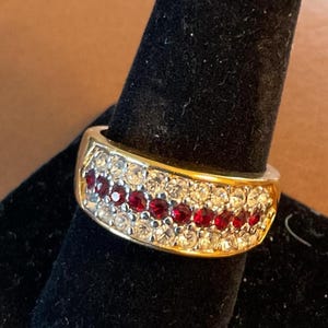 May include: A gold-toned ring with a band of clear and red gemstones. The ring is set on a black velvet surface, showcasing the sparkling stones. The gemstones are arranged in rows, creating a dazzling effect. A piece of jewelry.