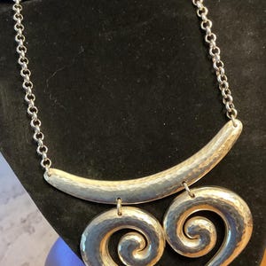 Puede incluir: Collar plateado con textura martillada. El collar presenta una barra curva en la parte superior, conectada a una cadena, y dos colgantes grandes en forma de coraz&oacute;n arremolinado. Se exhibe sobre una superficie de terciopelo negro.