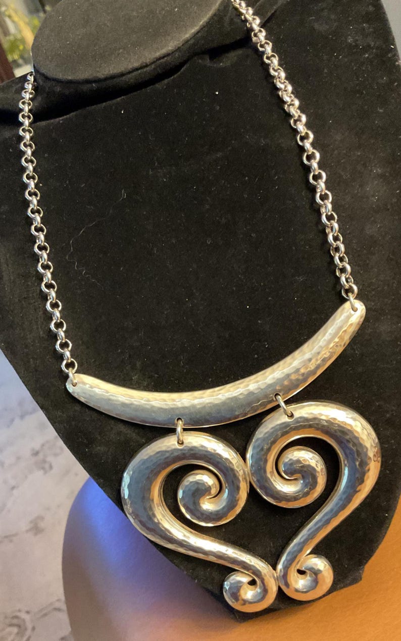Puede incluir: Collar plateado con textura martillada. El collar presenta una barra curva en la parte superior, conectada a una cadena, y dos colgantes grandes en forma de coraz&oacute;n arremolinado. Se exhibe sobre una superficie de terciopelo negro.