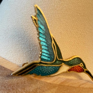 May include: A gold-toned hummingbird brooch with teal and green enamel wings, a red throat, and a black beak. The brooch is detailed with white and gold accents, showcasing a vibrant and elegant design.
