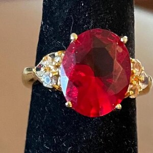 May include: A gold-toned ring featuring a large, oval-cut, red gemstone as the centerpiece. The ring is accented with smaller, clear gemstones on either side of the main stone. The ring is displayed on a black surface.