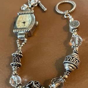 May include: A silver-toned watch bracelet featuring a rectangular watch face with a white dial and black hands. The band consists of alternating clear faceted glass beads and ornate silver-toned metal beads with intricate patterns and a toggle clasp.