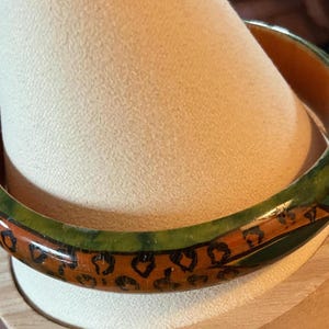 May include: A round bangle bracelet with a green and brown design. The bracelet has a green outer edge and a brown center with a leopard print pattern. The bracelet is displayed on a beige surface.