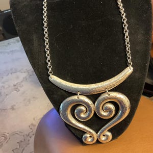 Puede incluir: Collar plateado con acabado martillado. El collar presenta una barra curva en la parte superior, conectada a una cadena, y dos colgantes grandes en forma de coraz&oacute;n con un dise&ntilde;o en espiral. El collar se exhibe sobre un busto de terciopelo negro.