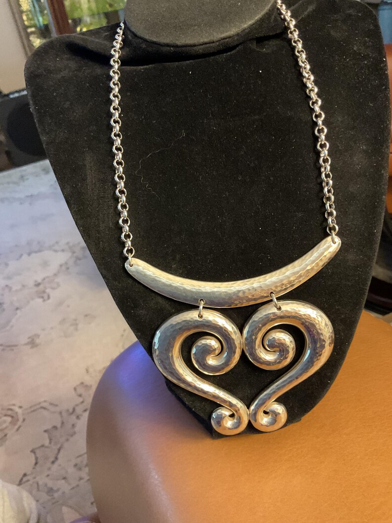Puede incluir: Collar plateado con acabado martillado. El collar presenta una barra curva en la parte superior, conectada a una cadena, y dos colgantes grandes en forma de coraz&oacute;n con un dise&ntilde;o en espiral. El collar se exhibe sobre un busto de terciopelo negro.