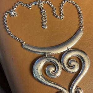 Puede incluir: Collar plateado con textura martillada. Presenta una barra curva y una cadena, con dos grandes colgantes en forma de coraz&oacute;n arremolinado. La cadena tiene un dije en forma de coraz&oacute;n cerca del cierre.