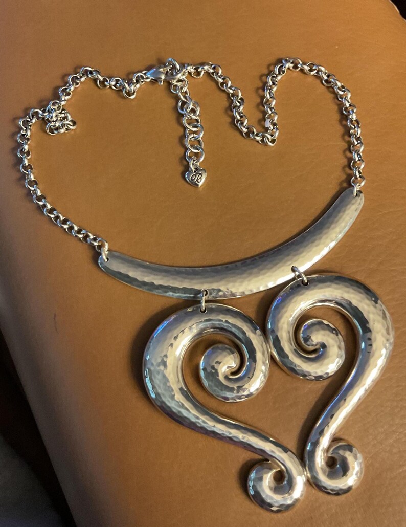 Puede incluir: Collar plateado con textura martillada. Presenta una barra curva y una cadena, con dos grandes colgantes en forma de coraz&oacute;n arremolinado. La cadena tiene un dije en forma de coraz&oacute;n cerca del cierre.