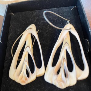 May include: A pair of cream-colored earrings with a unique, openwork design. The earrings have a layered, petal-like shape with subtle brown and beige accents. They are suspended from silver-toned ear wires, creating a striking and elegant accessory.