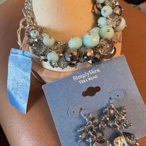 May include: A silver-toned bracelet and matching earrings with a cluster of beads in shades of gray and light blue. The earrings and bracelet are displayed on a neutral-colored stand. The Simply Vera Vera Wang brand is visible.