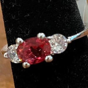 May include: A silver-toned ring featuring a central oval-cut red gemstone flanked by two smaller, round-cut clear stones. The stones are held in place by metal prongs on a thin, polished metallic band that wraps around a dark, textured display.