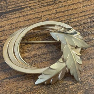 May include: A gold-toned brooch with a leaf design. The brooch features a circular shape with layered bands and a cluster of detailed leaves on one side. The pin is secured with a straight bar and clasp.