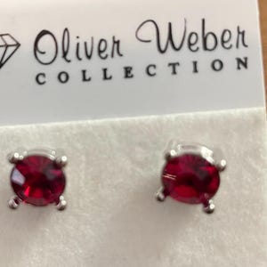 May include: A pair of stud earrings with round, deep red gemstones set in silver-colored metal. The earrings are displayed on a white card with the text "Oliver Weber COLLECTION" printed in black.