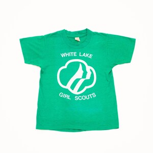 K&ouml;nnte beinhalten: Ein gr&uuml;nes T-Shirt mit dem wei&szlig;en Schriftzug "WHITE LAKE GIRL SCOUTS". Das Design zeigt eine wei&szlig;e Umrandung von zwei Profilen in einer Wolkenform. Das Shirt besteht aus einem weichen Material.