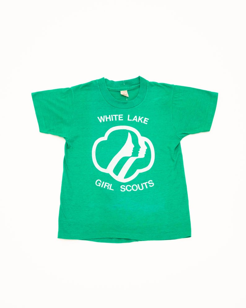 K&ouml;nnte beinhalten: Ein gr&uuml;nes T-Shirt mit dem wei&szlig;en Schriftzug "WHITE LAKE GIRL SCOUTS". Das Design zeigt eine wei&szlig;e Umrandung von zwei Profilen in einer Wolkenform. Das Shirt besteht aus einem weichen Material.