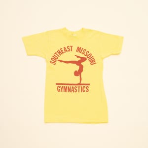 Puede incluir: Camiseta amarilla de manga corta con un gráfico rojo de una gimnasta en una viga de equilibrio. El texto "Southeast Missouri Gymnastics" está impreso en rojo alrededor del gráfico.