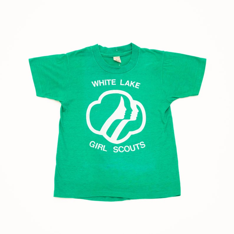 K&ouml;nnte beinhalten: Ein gr&uuml;nes T-Shirt mit dem wei&szlig;en Text "WHITE LAKE GIRL SCOUTS". Das Design zeigt eine wolkenartige Form mit zwei Silhouetten. Das Shirt hat einen Rundhalsausschnitt und kurze &Auml;rmel.