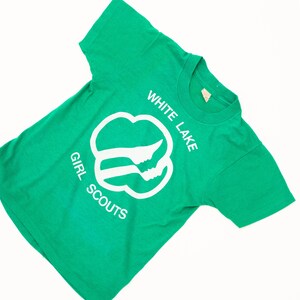 K&ouml;nnte beinhalten: Ein gr&uuml;nes T-Shirt mit den wei&szlig;en Worten "WHITE LAKE GIRL SCOUTS". Das Shirt hat ein wei&szlig;es Emblem mit zwei Silhouetten. Das T-Shirt ist aus einem weichen Material gefertigt.