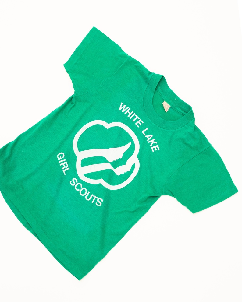 K&ouml;nnte beinhalten: Ein gr&uuml;nes T-Shirt mit den wei&szlig;en Worten "WHITE LAKE GIRL SCOUTS". Das Shirt hat ein wei&szlig;es Emblem mit zwei Silhouetten. Das T-Shirt ist aus einem weichen Material gefertigt.