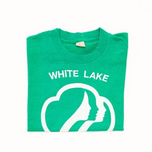 K&ouml;nnte beinhalten: Ein gefaltetes gr&uuml;nes T-Shirt mit dem wei&szlig;en Text "WHITE LAKE" &uuml;ber einer wei&szlig;en Grafik von zwei Frauenprofilen in einer Wolkenform. Das T-Shirt ist aus einem weichen Material.