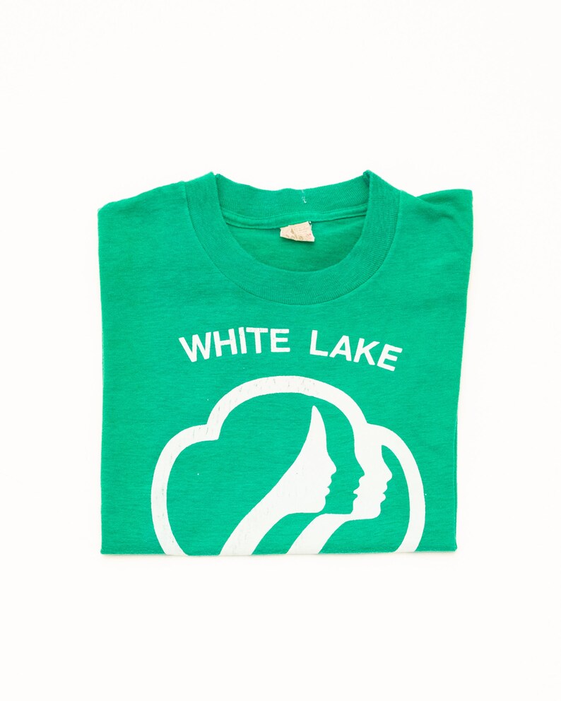 K&ouml;nnte beinhalten: Ein gefaltetes gr&uuml;nes T-Shirt mit dem wei&szlig;en Text "WHITE LAKE" &uuml;ber einer wei&szlig;en Grafik von zwei Frauenprofilen in einer Wolkenform. Das T-Shirt ist aus einem weichen Material.