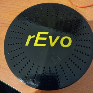 Könnte beinhalten: Eine schwarze kreisförmige Scheibe mit einem gelben "rEvo"-Logo und einem Muster aus kleinen Löchern.