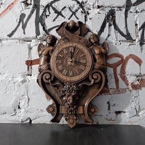 Puede incluir: Un reloj de pared de madera con un diseño tallado. La esfera del reloj tiene números romanos y está flanqueada por dos figuras talladas. Debajo de la esfera del reloj hay una cara tallada con cuernos. El reloj está montado en una pared de ladrillo blanco con graffiti.
