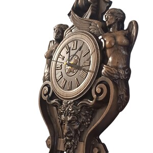 May include: A bronze-colored wall clock with Roman numerals and ornate carvings. The clock features two sculpted figures, wings, and a face with a beard. The clock hands point to approximately 2:10.