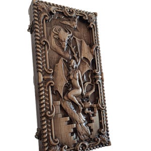 May include: A rectangular wooden box featuring an intricate relief carving on its lid. The design depicts a winged figure seated on a stone structure beneath a large horned creature with expansive wings, all framed by a decorative rope-like border.