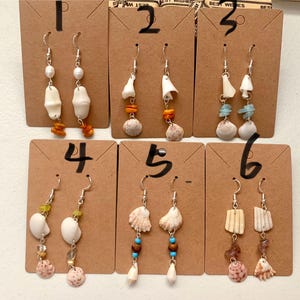 May include: Six pairs of dangle earrings displayed on brown cardboard cards numbered one through six. Each pair features silver-toned hooks with various combinations of white seashells, small colourful beads, and natural stones in diverse shapes and sizes.