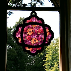 Könnte beinhalten: Ein schwarzes Ornament mit floralem Muster und einem gewellten Rand hängt an einem Fenster. Das Ornament ist aus Papier oder Stoff gefertigt und zeigt eine Vielzahl von rosa, roten und orangefarbenen Blumen.