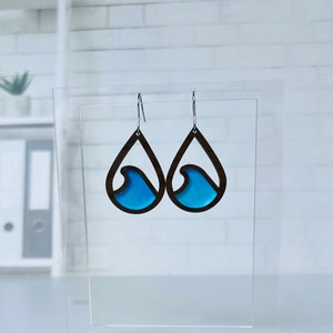May include: A pair of teardrop-shaped wooden earrings with a blue wave design. The earrings are hanging from silver hooks.