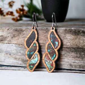 May include: A pair of long, dangle earrings with a wood and resin design. The earrings are made of three overlapping pieces of wood, each with a different pattern. The wood is a light brown color, and the resin is a mix of blue, green, and brown colors.