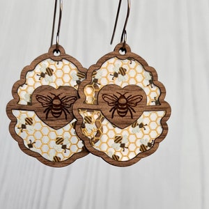 May include: A pair of wooden earrings with a honeycomb pattern and a bee in a heart design. The earrings are brown and have silver hooks.