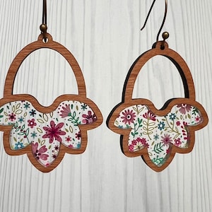 Puede incluir: Un par de pendientes de madera con un patrón floral. Los pendientes tienen forma de campana y un diseño recortado. El patrón floral es en tonos de rosa, blanco y verde.