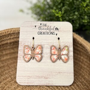 Peut inclure: Une paire de boucles d'oreilles en bois en forme de papillon avec un motif floral. Les boucles d'oreilles sont sur une carte d'affichage en bois avec le texte "The Thankful Creations" imprimé dessus.