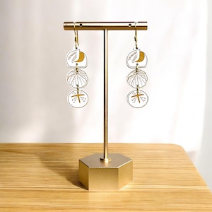 May include: Gold-colored earring display stand with three pairs of white and gold earrings. Each earring features a layered design with a crescent moon, a sunburst, and a cross-like shape. The earrings are hanging on a gold-colored stand.