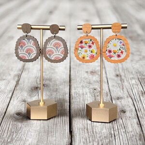 May include: Two pairs of wooden earrings on a gold stand. The earrings are round and have a scalloped edge. One pair features a floral pattern and the other pair features a geometric pattern.