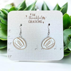 May include: A pair of dangle earrings featuring a white oval design with a brown wheat stalk and the word "SOURDOUGH". The earrings are suspended from silver-colored hooks. The earrings are displayed on a gray card with the text "THE thankful CREATIONS".