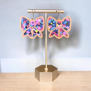 May include: A pair of wooden bow earrings with a colorful abstract pattern. The earrings are displayed on a gold stand.