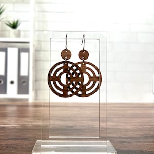 Pode incluir: Um par de brincos de madeira com um design geométrico. Os brincos são marrons e têm uma forma circular com um padrão de círculos concêntricos.