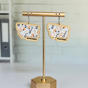 May include: A pair of wooden earrings shaped like the state of Montana, with a floral pattern in shades of orange, brown, and white. The earrings are hanging from a gold stand.