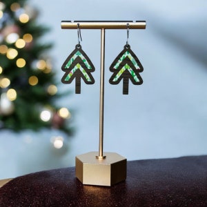May include: A pair of wooden Christmas tree earrings with a green and white floral pattern. The earrings are displayed on a gold stand.