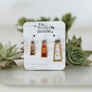 May include: Three pairs of wooden earrings with floral designs. The earrings are displayed on a white wooden board with the text "THE thankful CREATIONS" printed on it.